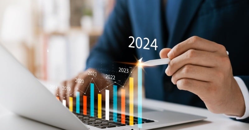 person in suit seated at desk in front of laptop multicolored hologram bar graph indicating revenue growth by year ending in 2024 stylus pointed at last bar graph