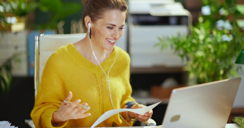 woman in yellow sweater with earbuds holding a piece of paper smiling seated at desk looking at computer office plants in background