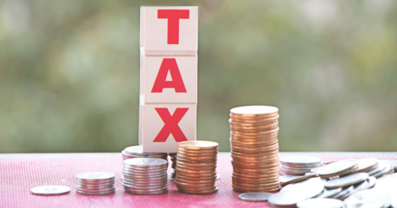 white domino with red ledders spelling "TAX" veritcally on top of stacks of coins in different denominations on pink table with sage green background