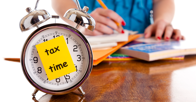 Alarm clock with yellow sticky note that says "Tax Time" on clock face sitting on brown desk in front of person with red nail polish and blue blouse working on tax forms in background