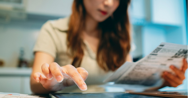 woman in white shirt reading from a paper while seated at desk her finger hovering over a tablet in the foreground